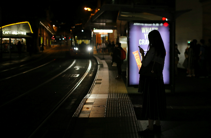 A woman uses her phone while standing in front of an illuminated advertisement as a train approaches in central Sydney, Australia.